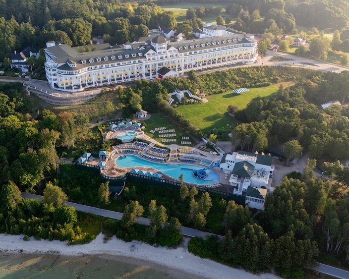 Aerial view of a large white hotel with multiple wings, surrounded by trees and a sprawling pool area with lounge chairs and water features near the shore.
