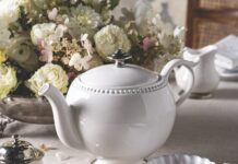 Before She Says “I Do” White porcelain teapot on a light table with a bouquet of white flowers in the background.