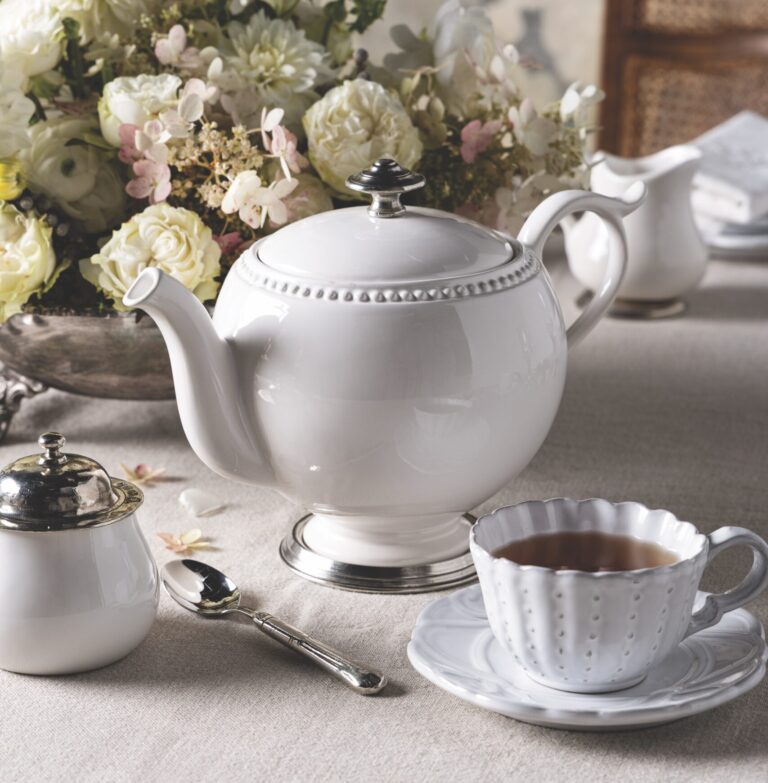 White porcelain teapot on a light table with a bouquet of white flowers in the background.