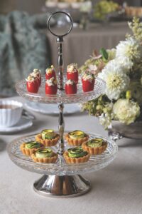 Two-tier crystal serving stand filled with red pepper canapé bites topped with cheese, set beside a bouquet of white flowers.