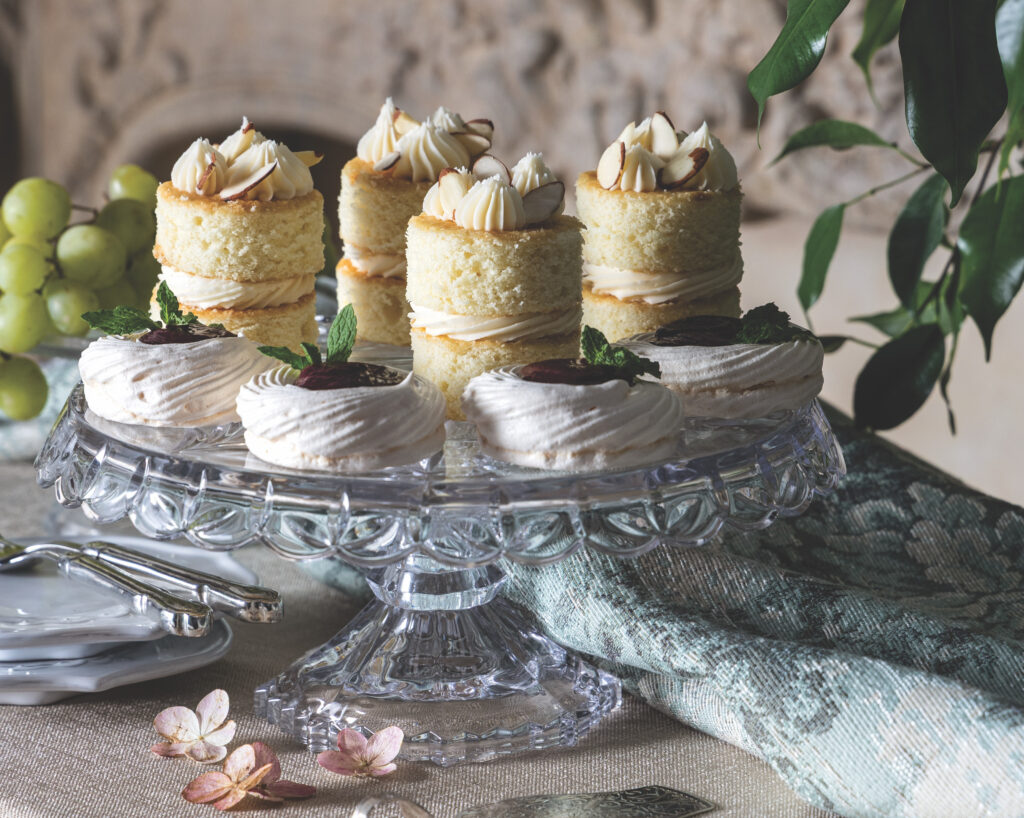 Assorted mini cakes with cream frosting and almond topping on a glass stand, surrounded by grapes and greenery.