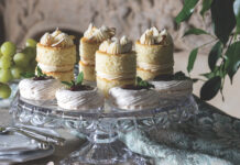 Petite Lemon Pavlovas with Blueberry Curd Assorted mini cakes with cream frosting and almond topping on a glass stand, surrounded by grapes and greenery.