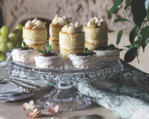 Assorted mini cakes with cream frosting and almond topping on a glass stand, surrounded by grapes and greenery.