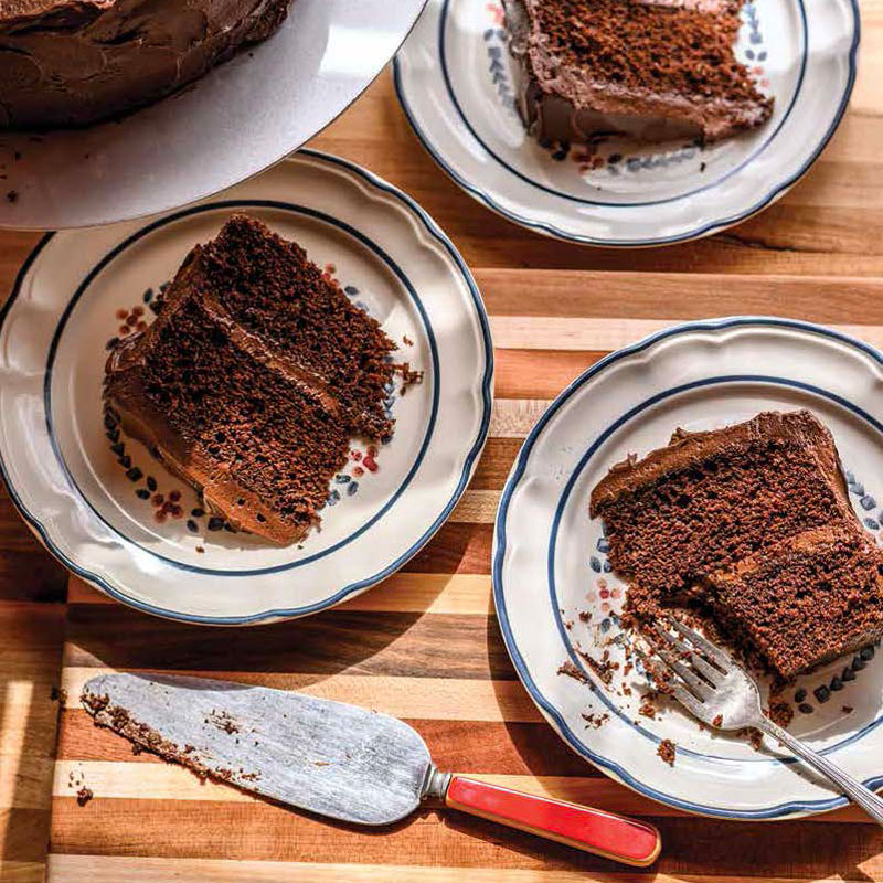 Slices of chocolate layer cake on ceramic plates with a wooden table and a red-handled cake server nearby
