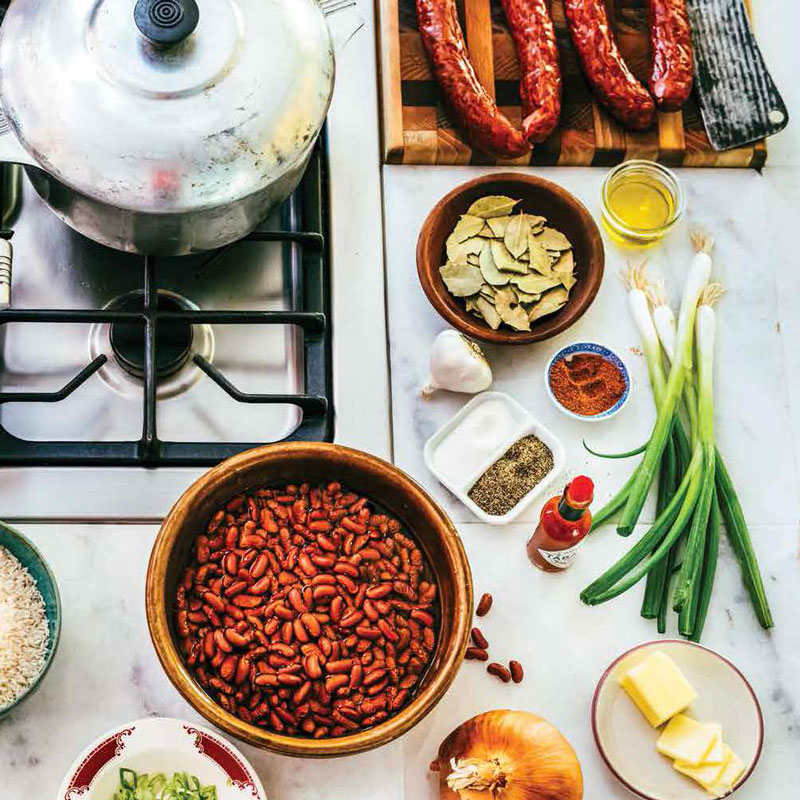 Top-down view of a stovetop and prep area with sausages, a pot, and bowls of beans, onions, green onions, garlic, spices, and butter ready for cooking.