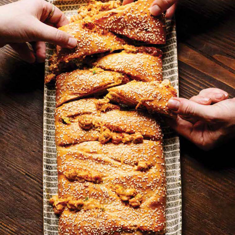 Hands pull apart a sesame-seed crusted loaf of bread on a patterned platter.