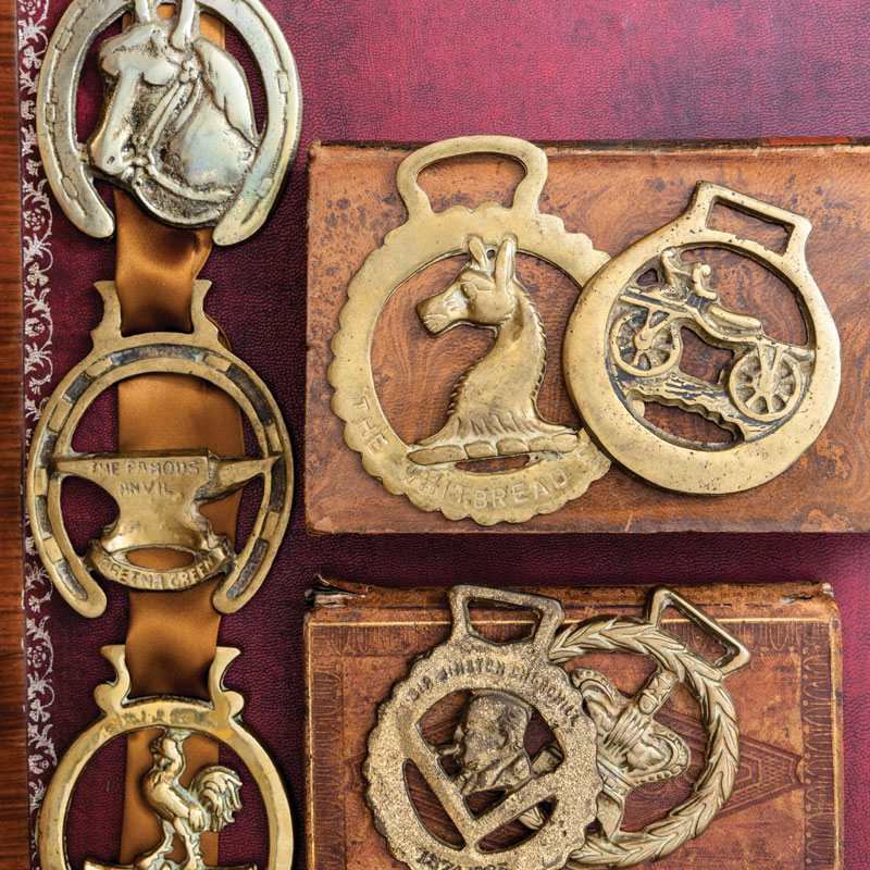 Set of antique brass belt buckles and medallions laid on a burgundy surface, featuring horse and animal emblems, a bicycle cutout, and laurel motifs.