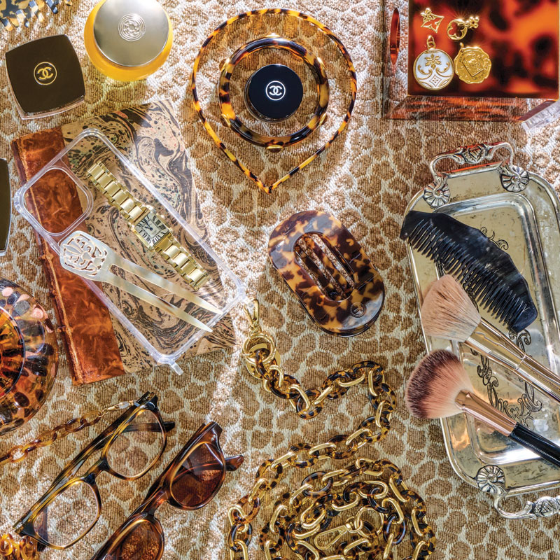 Assorted luxury accessories on patterned fabric: Chanel items, gold chains, tortoiseshell clips, brushes, and sunglasses laid out.