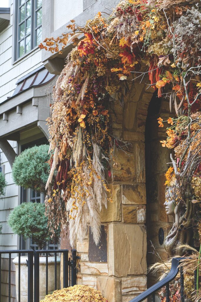 A close-up of a stone doorframe arch covered in twining branches, dried hydrangea, pampas grass, and autumn foliage in rust and gold.