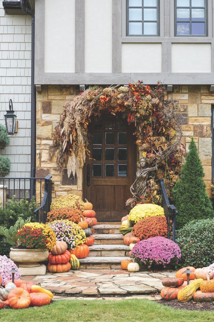 A wooden front door framed by a sculptural installation of twining branches, pampas grass, and dried florals above steps lined with mums and pumpkins.
