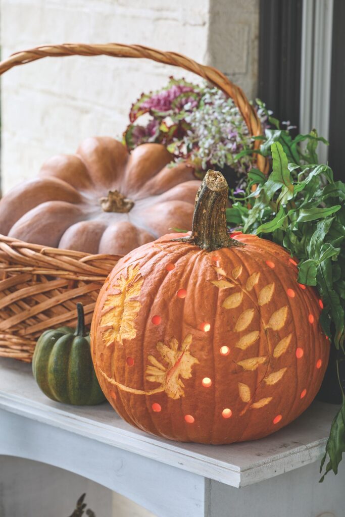 An orange pumpkin carved with a delicate leaf pattern sits beside a woven basket holding a large tan pumpkin and dried hydrangeas.