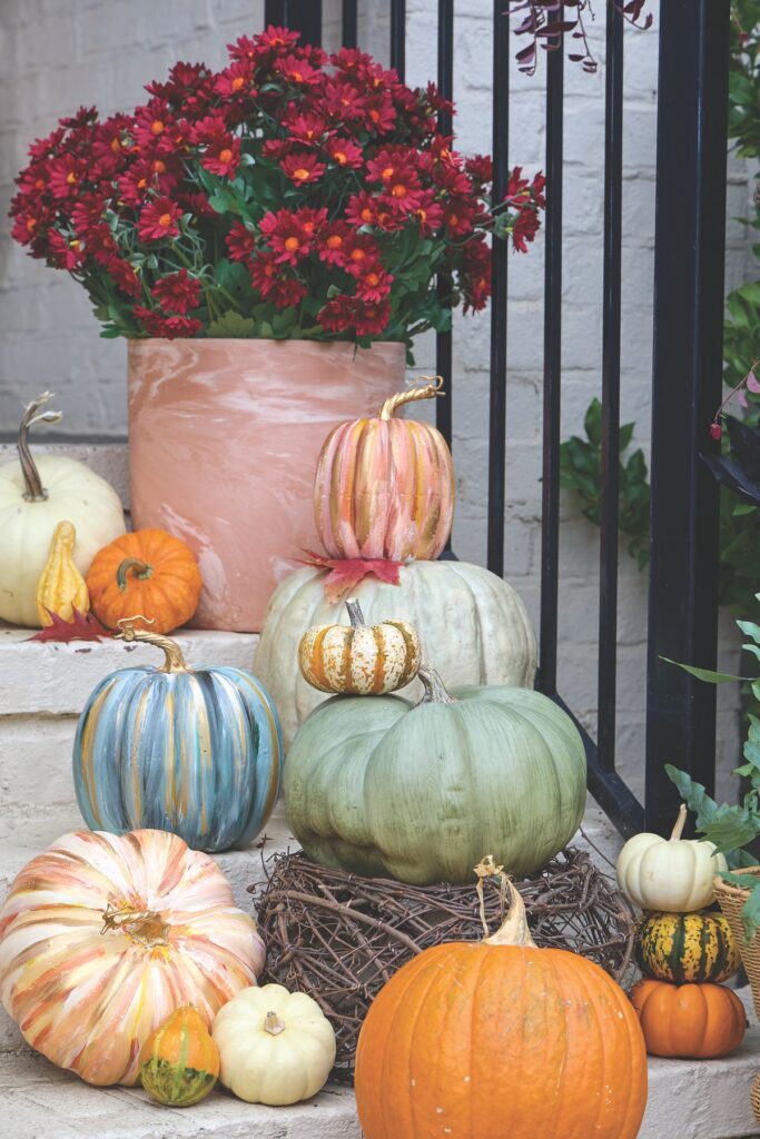 Pumpkins painted with streaks of blue, green, peach, and gold stacked on porch steps beneath a pot of deep red mums.