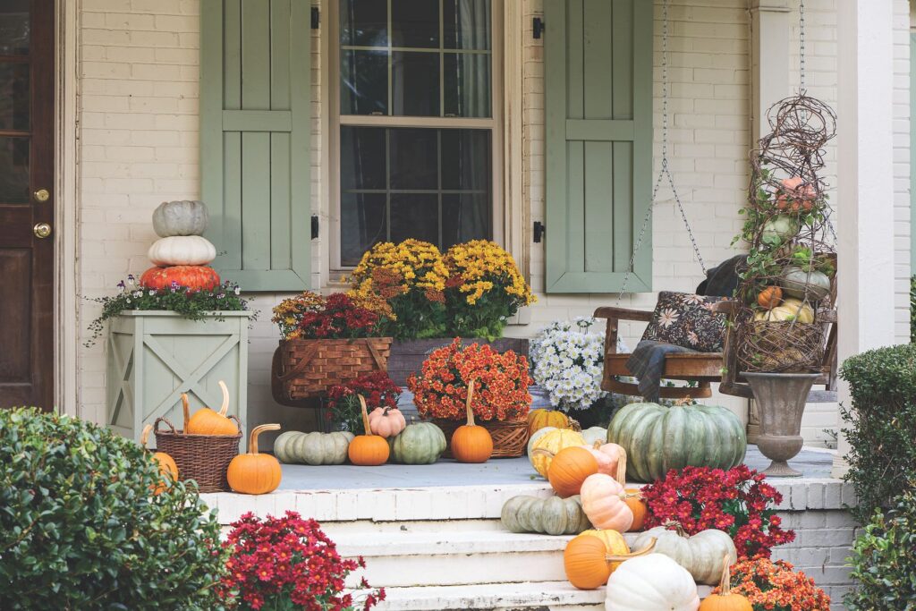 A white brick porch with sage green shutters styled with a planter box, stacked pastel pumpkins, and baskets and urns of jewel-toned mums.
