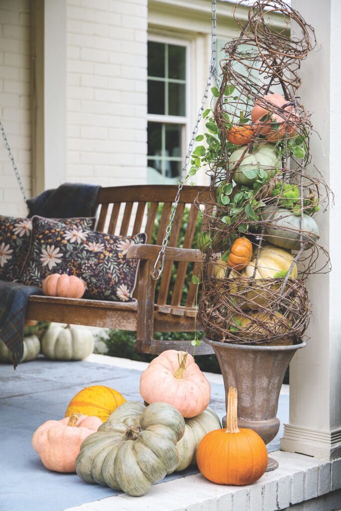 A wooden porch swing with patterned pillows beside an urn topped with a grapevine sphere and pastel pink, green, and orange pumpkins.