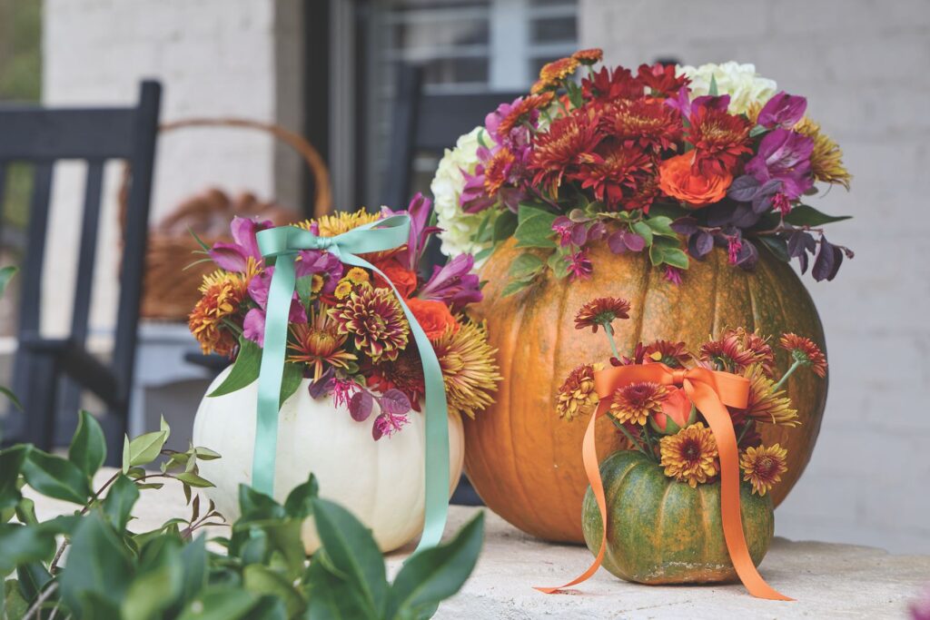 Pumpkins used as vases for arrangements of red, orange, and purple autumn blooms, tied with teal and orange ribbons.