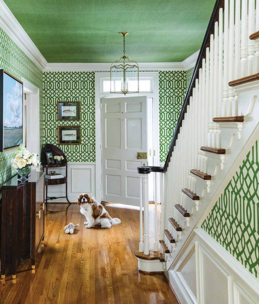 A green trellis-papered entryway with a lacquered green ceiling, white staircase, and a spaniel resting on the wood floor