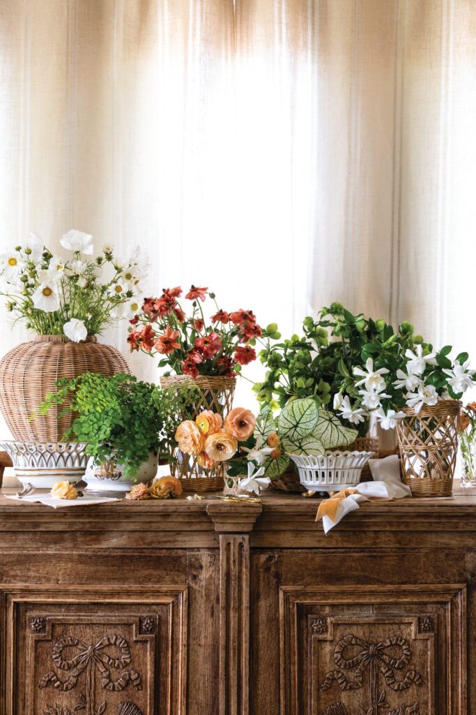 Ranunculus, cosmos and gardenias arranged in wicker and silver vessels on a carved wooden sideboard