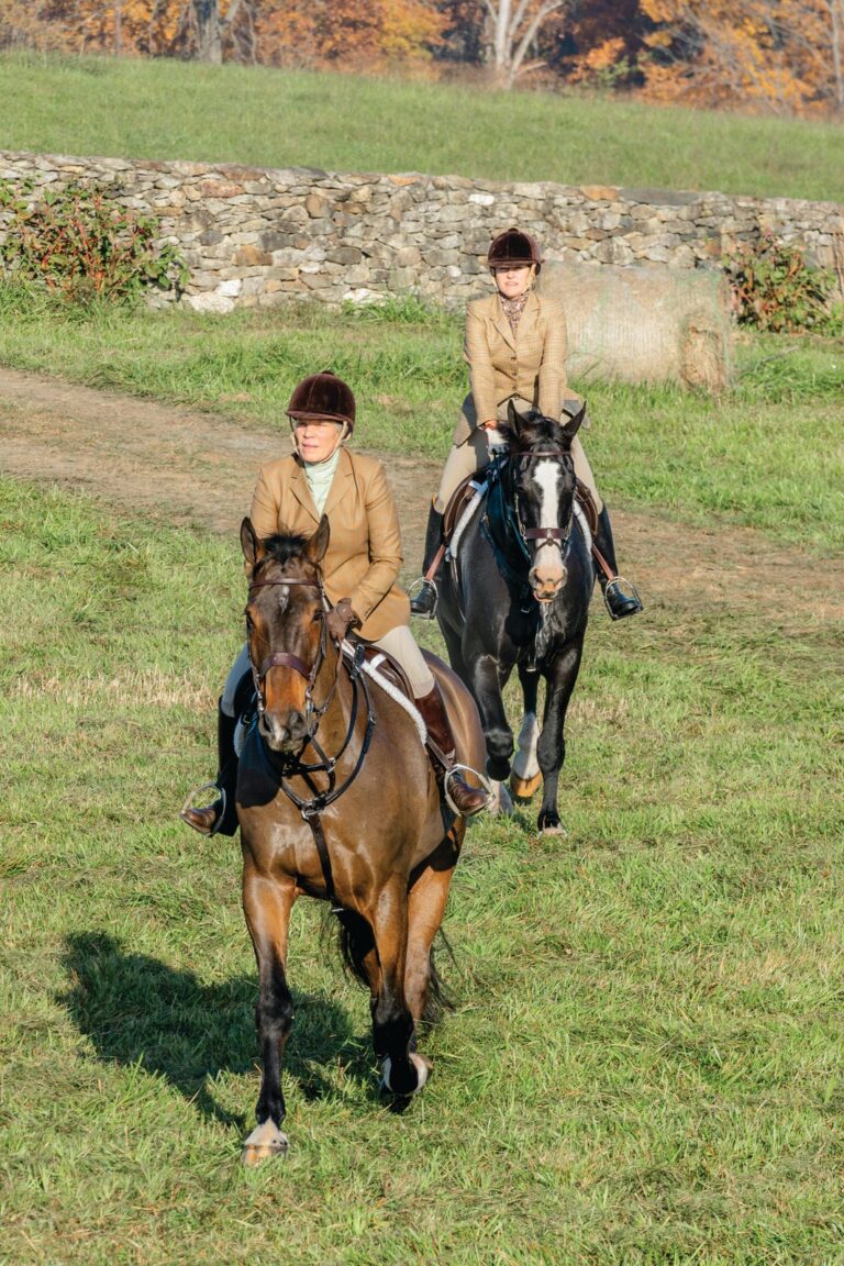 Two riders in hunt attire canter across an autumn field past a low stone wall in Middleburg, Virginia